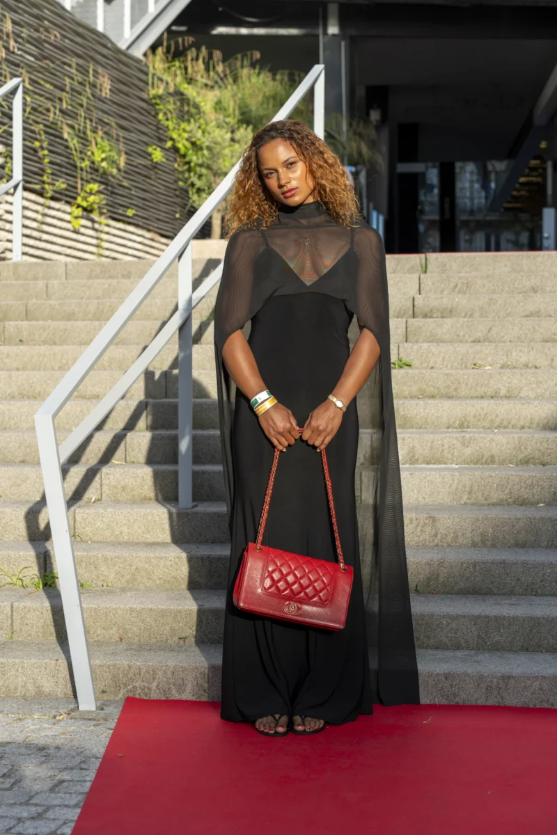 Cette photographie met en scène une femme comme sujet principal, debout sur un tapis rouge. Elle porte une robe noire élégante et des accessoires voyants, dont un sac à motifs colorés.La composition est symétrique, avec un cadrage serré sur elle, encadrée par des marches et une rampe. La lumière est naturelle et douce, mettant en valeur les contours.Les couleurs dominantes sont le noir, le rouge du tapis et les touches vives du sac. Le style est sophistiqué et chic, évoquant une ambiance de gala ou événement mondain.