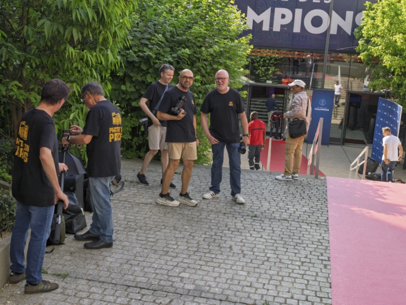 Thierry · 23/06/2026 · 3840 x 2560 Groupe de personnes en t-shirts noirs discutant près d’un espace festif avec une bannière 'Les Des Champions', sous