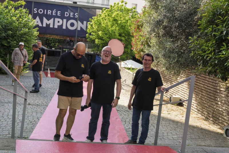 Thierry · 23/06/2026 · 3840 x 2560 Cette photographie montre trois hommes marchant sur un tapis rouge. Le sujet principal semble être un événement sportif, suggéré par les maillots noirs et les insignes, ainsi que l’enseigne "Soirée des Champions".La composition est symétrique, avec les hommes alignés sur le tapis. Le cadrage est serré, mettant en avant les personnages et le tapis rouge. La lumière est naturelle et douce, sans ombres marquées. Les couleurs dominantes sont le noir (vêtements), le rouge (tapis) et le vert (végétation en arrière-plan).Le style est documentaire, capturant un moment réel. L’ambiance est solennelle et festive, typique d’une cérémonie officielle.