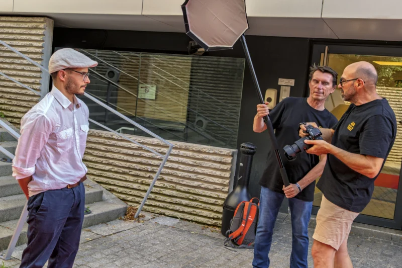 Thierry · 23/06/2026 · 3840 x 2560 Cette photographie montre trois hommes en extérieur, impliqués dans une activité de tournage ou d’interview. Le sujet principal semble être l’interaction entre eux, avec un homme tenant un micro et un autre manipulant une caméra.La composition est dynamique, avec un cadrage serré sur les visages et les mains. La lumière est naturelle, probablement en journée, éclairant les sujets de manière uniforme. Les couleurs dominantes sont neutres et sobres, avec des tons gris, beiges et quelques touches de couleurs vives sur les vêtements.L’ambiance est professionnelle et informelle, évoquant un tournage amateur ou semi-professionnel.