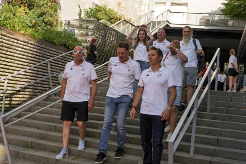 Thierry · 23/06/2026 · 3840 x 2560 Cette photographie montre un groupe de sept personnes posant sur un escalier extérieur. Le sujet principal est ce groupe, vêtu de tenues sportives blanches, avec des poses dynamiques et souriantes.La composition est symétrique, avec les individus alignés sur les marches. Le cadrage est serré, mettant en valeur les visages et les gestes. La lumière est naturelle, douce et légèrement tamisée, probablement en fin de journée. Les couleurs dominantes sont les tons clairs (blanc des vêtements, gris des marches) avec des touches de bleu et vert en arrière-plan.L’ambiance est conviviale et énergique, évoquant une équipe unie.