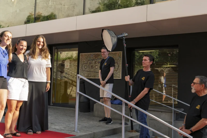 Thierry · 23/06/2026 · 3840 x 2560 Cette photographie montre un groupe de cinq personnes lors d’un événement, probablement une cérémonie ou une interview. Le sujet principal semble être l’homme tenant un micro, au centre, entouré des autres.La composition est symétrique avec un escalier menant à une entrée. Le cadrage est serré sur les personnes, avec un arrière-plan architectural sobre. La lumière est naturelle et douce, sans ombres marquées. Les couleurs dominantes sont neutres (gris, beige) avec des touches de bleu et vert.L’ambiance est formelle et professionnelle, évoquant un moment officiel ou médiatique.
