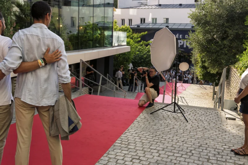 Thierry · 23/06/2026 · 3840 x 2560 Cette photographie montre une scène de tournage ou d'événement. Le sujet principal est un homme agenouillé près d’un réflecteur, probablement un technicien ou un photographe, tandis qu’un autre homme, de dos, semble discuter ou observer.La composition inclut une allée rouge menant à un escalier, encadrée par des bâtiments et des arbres. Le cadrage est serré sur les personnages et l’équipement, avec une profondeur de champ limitée.Les couleurs dominantes sont le rouge vif de la moquette, les tons neutres des vêtements et le vert des arbres. La lumière est naturelle, avec des ombres douces. L’ambiance est professionnelle et dynamique, évoquant un événement organisé.