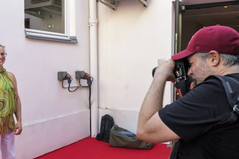 Thierry · 23/06/2026 · 3840 x 2560 Cette photographie montre un photographe en train de prendre une photo d’une personne vêtue d’une robe verte, partiellement visible dans un cadre de porte.La composition est simple, avec un cadrage serré sur le photographe et un arrière-plan minimaliste. La lumière semble naturelle, bien répartie, sans ombres marquées. Les couleurs dominantes sont les tons verts de la robe, le rouge du tapis et les teintes neutres des murs.Le style est documentaire et spontané, évoquant une ambiance authentique et décontractée, presque théâtrale.