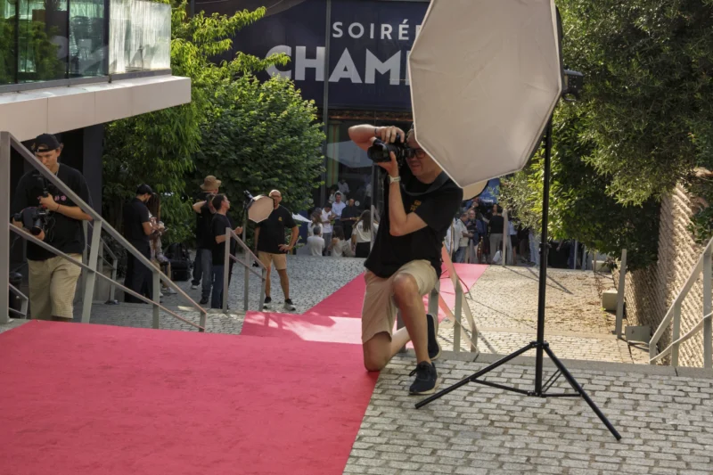 Thierry · 23/06/2026 · 3840 x 2560 Cette photographie capture une scène de soirée chic, probablement un événement lié au champagne. Le sujet principal est une personne en robe noire, filmée de dos, marchant sur un tapis rouge. La composition met en valeur le tapis rouge contrastant avec le sol pavé clair.Le cadrage est serré sur la personne, avec une lumière naturelle et artificielle douce. Les couleurs dominantes sont le rouge du tapis, le noir de la robe et les tons neutres des pavés et des lumières. L’ambiance est élégante et sophistiquée, typique d’un événement mondain.