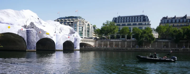 Bao · 5472 x 2143 Cette photographie montre un pont parisien avec des sculptures architecturales blanches en forme d’arcs, couvrant partiellement la Seine. Le sujet principal est ce pont temporaire, contrastant avec les bâtiments classiques en arrière-plan.La composition est horizontale, avec un cadrage large incluant une partie du fleuve et un bateau à droite. La lumière est naturelle et claire, évoquant une journée ensoleillée. Les couleurs dominantes sont les tons gris-beige des bâtiments, le bleu de l’eau et le blanc des structures.Le style est documentaire et urbain, créant une ambiance à la fois moderne et historique.