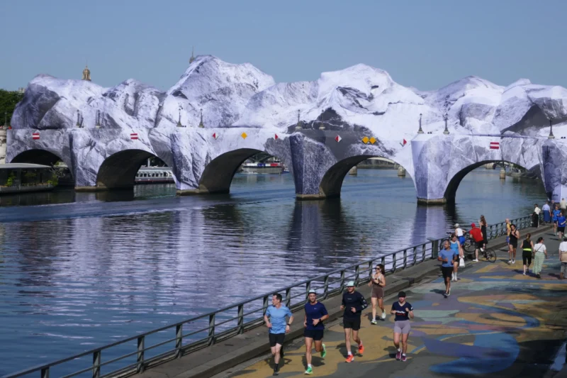 Bao · 5472 x 3648 Cette photographie montre un pont recouvert d’une installation artistique représentant des montagnes enneigées. Le sujet principal est cette structure transformée, créant un contraste saisissant avec l’eau calme en contrebas.La composition est symétrique, avec le pont au centre et des personnes en mouvement sur les trottoirs. Le cadrage inclut une partie du rivage et du ciel dégagé. La lumière est naturelle et vive, accentuant les détails des sculptures.Les couleurs dominantes sont les blancs éclatants des montagnes artificielles, contrastant avec le bleu de l’eau et le gris des structures. L’ambiance est à la fois ludique et artistique, évoquant une fusion entre nature et création humaine.