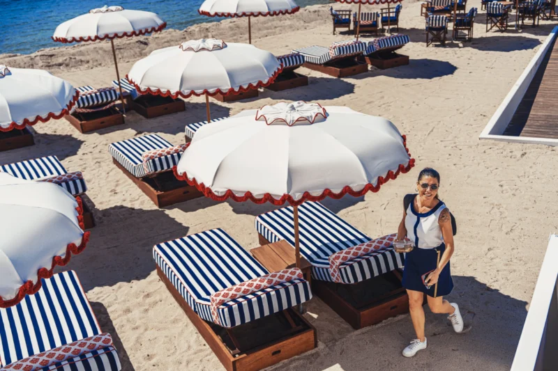 Marc L. · 13/05/2025 · 9504 x 6336 Cette photographie met en scène une femme comme sujet principal, vêtue d’une tenue sportive, marchant sur une plage. La composition montre des transats bleus et blancs sous des parasols à rayures rouges et blanches. Le cadrage est serré sur la personne, avec un arrière-plan légèrement flou mais identifiable.La lumière est vive et naturelle, typique d’un jour ensoleillé. Les couleurs dominantes sont les teintes claires (blanc, bleu) et les contrastes vifs des rayures rouges et blanches des parasols. Le style est dynamique et estival, évoquant détente et vacances. L’ambiance est joyeuse et relaxante, renforcée par le cadre balnéaire.