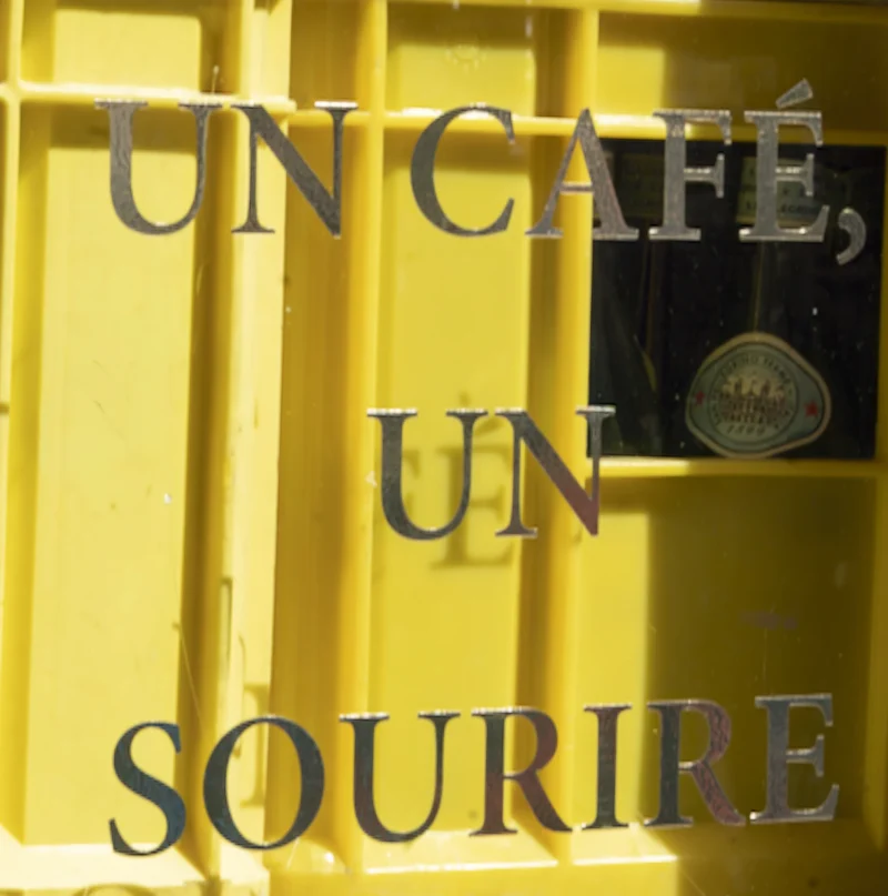 Jean-Yves · 20/06/2026 · 2975 x 3000 Cette photographie montre une enseigne vintage avec l'inscription "Un café, un sourire" sur un fond jaune pâle. Le sujet principal est le texte stylisé, encadré par une grille métallique.La composition est centrée, avec un cadrage serré sur l'enseigne. La lumière semble naturelle, douce et légèrement tamisée, créant des ombres subtiles. Les couleurs dominantes sont le jaune vif et le noir des lettres.Le style évoque un charme rétro et artisanal, renforçant une ambiance chaleureuse et accueillante, typique d'un café convivial.