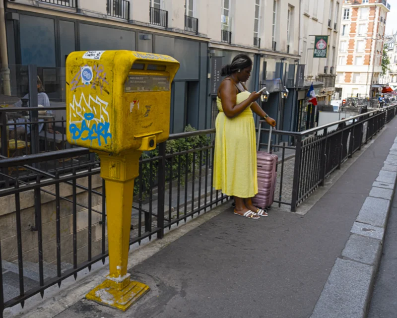 Jean-Yves · 20/06/2026 · 3000 x 2405 Cette photographie met en scène une femme en robe jaune clair comme sujet principal, posant devant un distributeur de tickets jaune décoré de motifs. La composition est simple et équilibrée, avec un cadrage serré sur la femme et l'objet.La lumière semble naturelle, probablement en journée, éclairant uniformément la scène. Les couleurs dominantes sont le jaune vif du distributeur et de la robe, contrastant avec les tons neutres de l'arrière-plan urbain. Le style est réaliste et documentaire, capturant un moment du quotidien. L'ambiance est urbaine et décontractée.