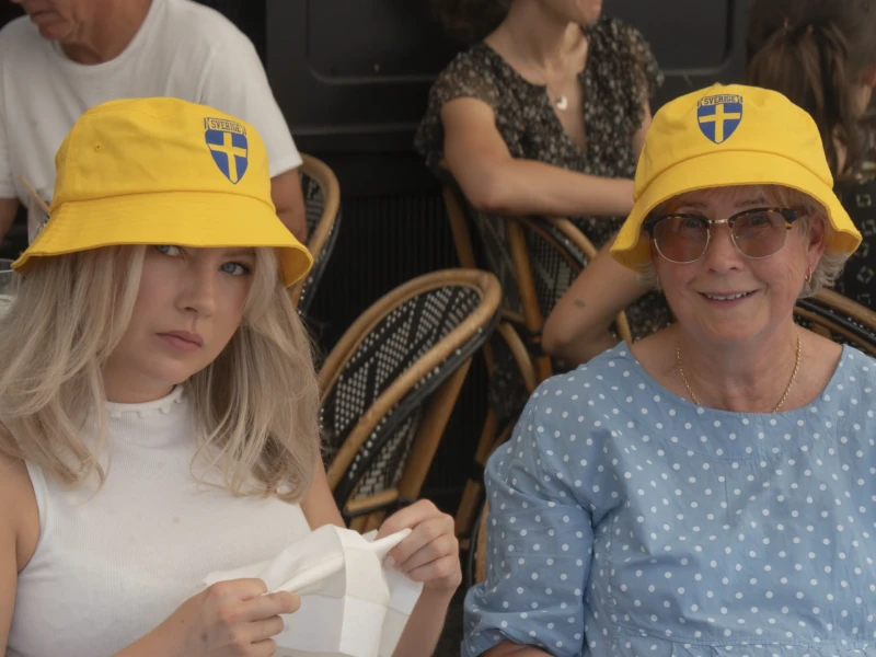 Jean-Yves · 20/06/2026 · 3000 x 2404 Cette photographie met en scène deux femmes portant des chapeaux jaunes à logo blanc. La jeune femme à gauche, vêtue d’un haut blanc, et la femme âgée à droite, en chemise bleue à pois, semblent attentives.Le cadrage est serré, centré sur elles, avec une personne floue en arrière-plan. La lumière est naturelle et douce, éclairant les visages. Les couleurs dominantes sont le jaune vif des chapeaux, le bleu de la chemise et le blanc du haut.L’ambiance est conviviale et détendue, évoquant un événement public ou une célébration. Le style est simple et spontané.