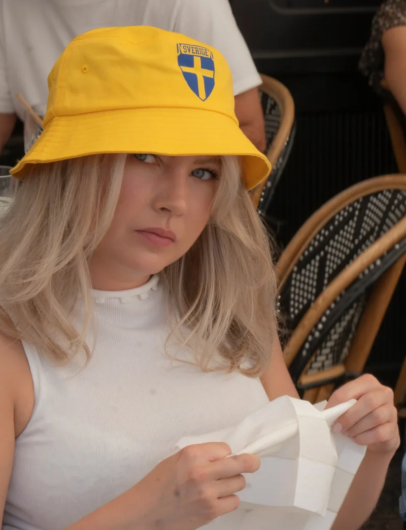 Jean-Yves · 1605 x 2097 Cette photographie montre une jeune femme comme sujet principal, assise et portant un chapeau jaune avec un emblème. Elle semble tenir un papier ou un objet blanc.La composition est simple, avec un cadrage serré sur son visage et ses épaules. L'arrière-plan est flou, mettant l'accent sur elle. La lumière est naturelle et douce, probablement en extérieur. Les couleurs dominantes sont le jaune vif du chapeau, le blanc de ses vêtements, et des teintes neutres pour le reste.Le style est naturel et spontané, évoquant une ambiance détendue, peut-être lors d’un événement en plein air.