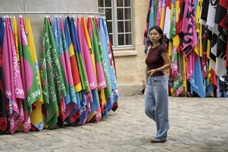 Thierry · 01/07/2026 · 3840 x 2560 Cette photographie met en scène une jeune femme comme sujet principal, marchant devant un étalage de foulards colorés. La composition est dynamique, avec un cadrage serré sur elle et une perspective légèrement basse. Les couleurs dominantes sont vives et variées (rose, bleu, jaune, vert), créant un contraste joyeux.La lumière semble naturelle, probablement en journée, éclairant les tons sans ombres marquées. L’ambiance est urbaine et vivante, évoquant un marché ou une rue animée. Le style est documentaire, capturant un moment authentique et coloré.