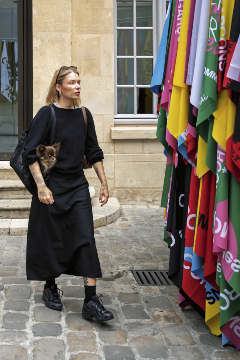 Thierry · 01/07/2026 · 2560 x 3840 Cette photographie met en scène une femme comme sujet principal, tenant un petit chien dans ses bras. La composition montre une rue pavée avec des étals de tissus colorés en arrière-plan.Le cadrage est serré sur la femme, avec une profondeur de champ qui floute légèrement les couleurs vives des tissus. La lumière est naturelle et douce, éclairant la scène sans créer d'ombres marquées. Les couleurs dominantes sont les tons chauds des tissus (rouge, jaune, vert) et les noirs de la tenue de la femme.Le style est réaliste et documentaire, capturant une ambiance urbaine et chaleureuse.