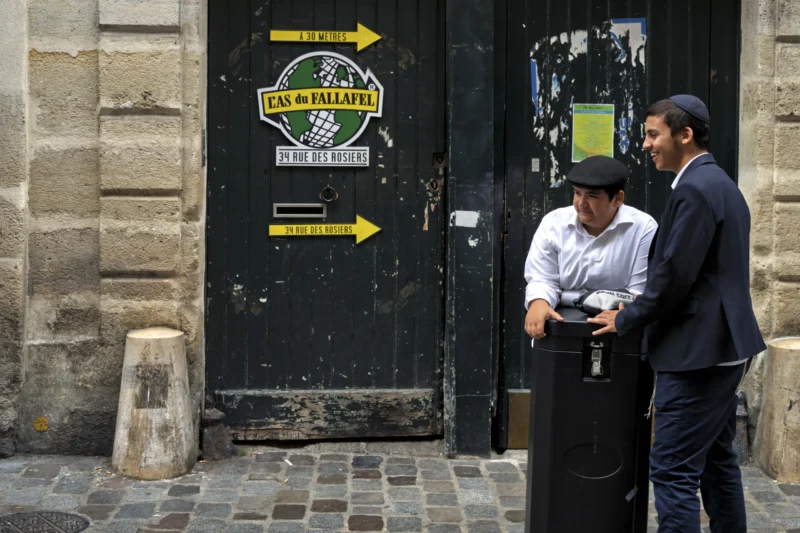 Thierry · 01/07/2026 · 3840 x 2560 Cette photographie montre deux hommes en costume discutant devant une porte en métal. Le sujet principal est leur échange, tandis que l'arrière-plan inclut une enseigne "Restaurant" avec des flèches directionnelles.Le cadrage est serré sur les deux personnages, avec une composition légèrement déséquilibrée vers la gauche. La lumière est naturelle, avec des ombres marquées, suggérant un éclairage diurne. Les couleurs dominantes sont les tons neutres (gris, noir) et quelques touches de vert et bleu sur les enseignes.Le style est réaliste et documentaire, capturant une scène urbaine du quotidien, créant une ambiance sobre et contemporaine.