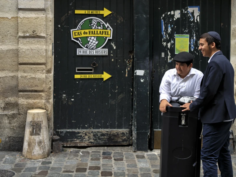 Thierry · 01/07/2026 · 3840 x 2560 Deux personnes discutent devant une porte noire avec une enseigne indiquant 'Le Falafel', sur un trottoir pavé en pierre.