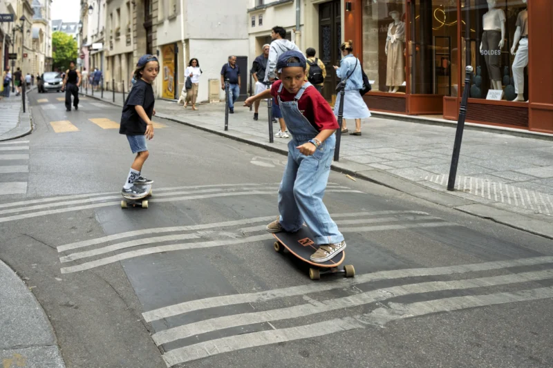 Thierry · 01/07/2026 · 3840 x 2560 Cette photographie capture un jeune homme comme sujet principal, en train de faire du skateboard sur une chaussée en pente. La composition montre une rue urbaine avec des passants en arrière-plan.Le cadrage est serré sur le skateur, mettant en valeur son action. La lumière est naturelle, légèrement tamisée, créant des ombres douces. Les couleurs dominantes sont les tons neutres (gris de la route, bleu du ciel) avec des éclats de rouge (vêtements) et vert (végétation).Le style est dynamique et spontané, évoquant une ambiance jeune et énergique dans un cadre urbain quotidien.