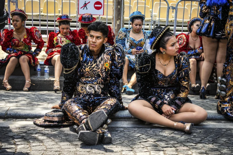 Thierry · 05/07/2026 · 3840 x 2560 Cette photographie capture deux personnes assises au premier plan, vêtues de costumes traditionnels ornés de motifs colorés et dorés. Leur posture et leurs expressions semblent contemplatives.La composition montre un groupe de personnes en arrière-plan, également habillées de manière similaire, créant une ambiance communautaire. Le cadrage est serré sur les deux sujets principaux, avec une profondeur de champ qui met l'accent sur eux.Les couleurs dominantes sont les noirs, ors et rouges, apportant une touche riche et vibrante. La lumière semble naturelle, éclairant les sujets de manière uniforme. L’ambiance est à la fois traditionnelle et solennelle, évoquant une célébration culturelle ou une cérémonie.