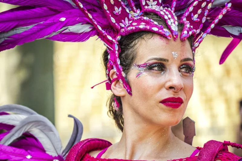 Thierry · 05/07/2026 · 3840 x 2560 Cette photographie met en scène une femme comme sujet principal, vêtue d’un costume extravagant. Elle porte un chapeau orné de plumes et de fleurs violettes, avec des éléments scintillants.Le cadrage est serré, centré sur son visage et son accessoire, isolant ainsi le sujet. La lumière est naturelle et met en valeur les détails colorés. Les couleurs dominantes sont le violet, le rose et le blanc, créant un effet festif et théâtral.Le style évoque le carnaval ou une célébration artistique. L’ambiance est joyeuse, exubérante et pleine de vie.