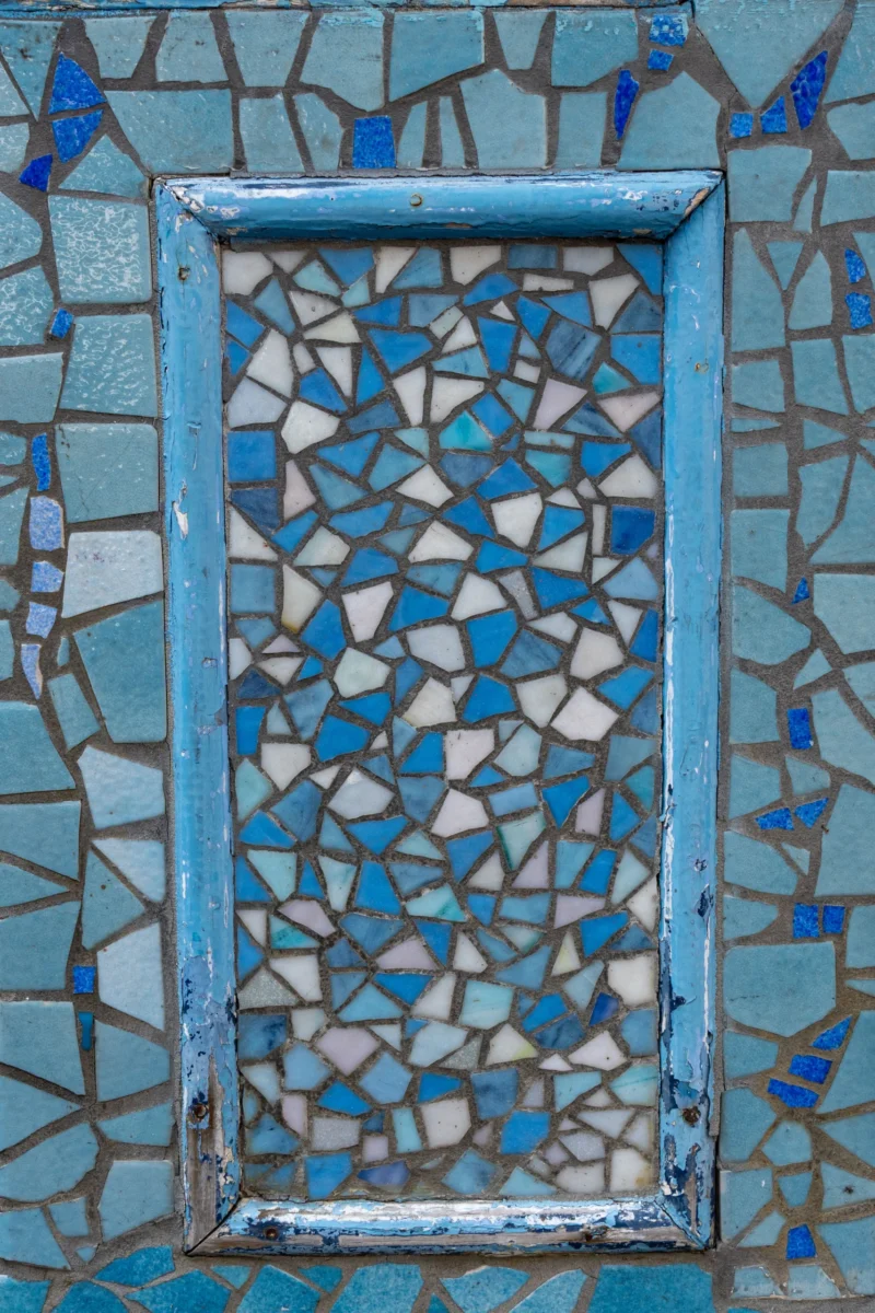 Marc L. · 20/06/2026 · 3539 x 5308 Cette photographie met en scène un cadre de fenêtre en métal peint en bleu délavé, encastré dans un mur recouvert de mosaïques aux motifs géométriques. Le cadrage est serré, centré sur le cadre et une partie du mur.Les couleurs dominantes sont le bleu et le blanc, avec des touches de gris. La lumière semble naturelle, éclairant le sujet de manière uniforme, créant des contrastes doux. Le style évoque l’artisanat traditionnel, mêlant simplicité et complexité des motifs. L’ambiance est à la fois rustique et artistique, avec une touche de nostalgie.