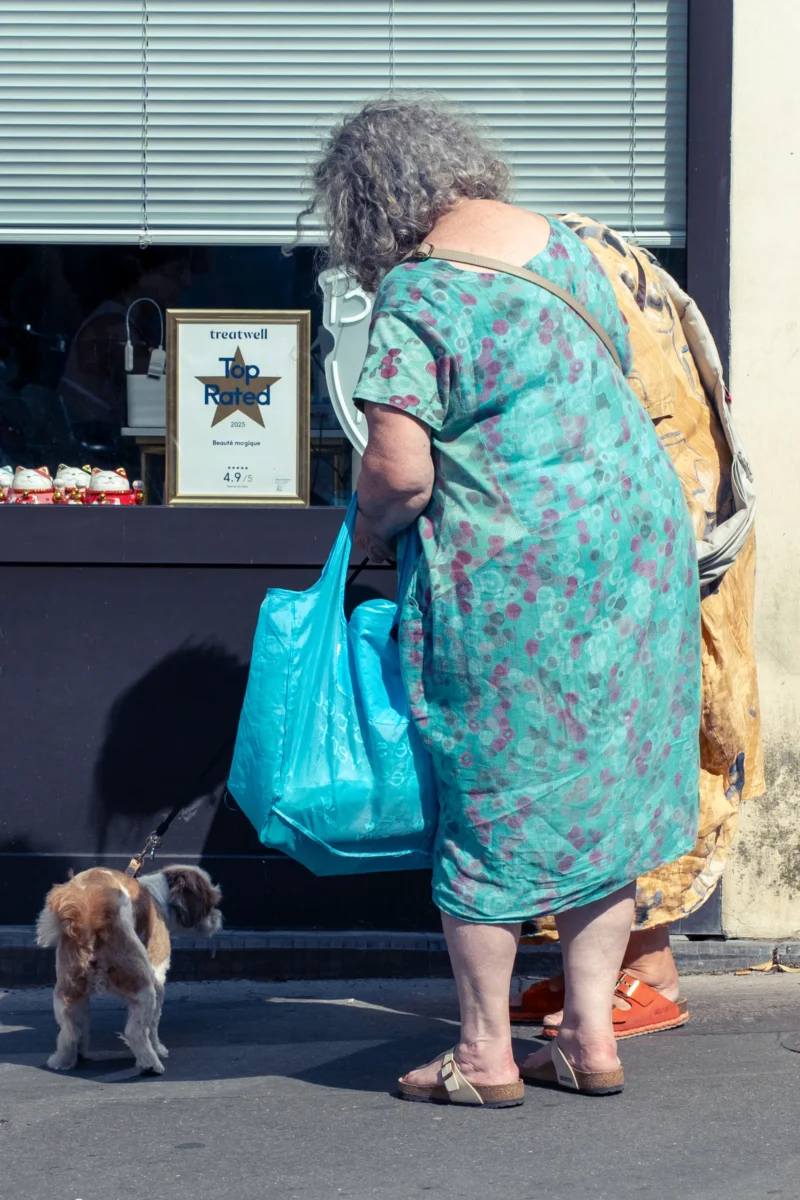 Marc L. · 20/06/2026 · 2131 x 3197 Cette photographie met en scène une femme âgée, sujet principal, vêtue d'une robe à motifs floraux, tenant un sac bleu. Elle est accompagnée d’un petit chien. La composition montre un cadrage serré sur la femme et son animal, avec un arrière-plan minimaliste incluant une vitrine et des objets décoratifs.La lumière est naturelle et douce, créant des ombres nettes. Les couleurs dominantes sont les teintes bleutées de la robe et du sac, contrastant avec les tons chauds de la peau et des accessoires. Le style est documentaire et spontané, évoquant une ambiance paisible et quotidienne.