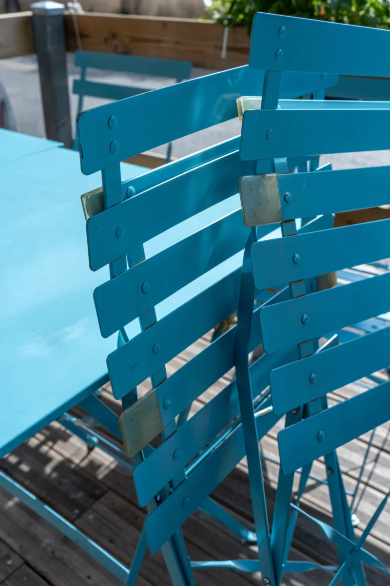 Marc L. · 20/06/2026 · 4672 x 7008 Cette photographie met en scène deux chaises pliante turquoise comme sujet principal. La composition est épurée, avec un cadrage serré sur les chaises, isolées sur un fond flou de terrasse en bois et de table bleue.La lumière semble naturelle, douce et légèrement tamisée, créant des ombres discrètes. Les couleurs dominantes sont le bleu turquoise vif des chaises et le bleu clair de la table, apportant une touche fraîche et moderne. Le style est minimaliste et contemporain, évoquant une ambiance apaisante et estivale, propice à la détente.