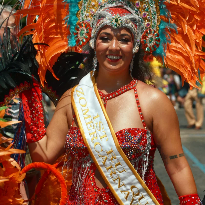 Jojo · 2656 x 2656 Cette photographie met en scène une femme en costume de reine de carnaval. Elle porte un sash doré avec l'inscription "Miss Carnaval", un costume rouge et or richement décoré, ainsi qu'un accessoire de tête coloré et festif.Le cadrage est serré, centré sur elle, avec des éléments de fond flous. La lumière est naturelle et met en valeur les couleurs vives. Les teintes dominantes sont le rouge, l'or et les tons chauds.Le style est festif et théâtral, évoquant une ambiance joyeuse et exubérante, typique des célébrations carnavalesques.