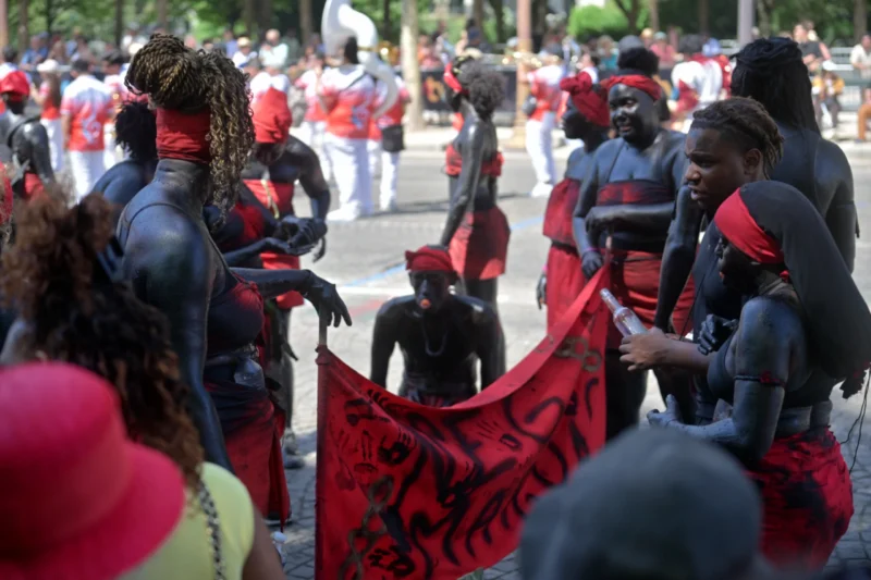 Jojo · 05/07/2026 · 4096 x 2730 Cette photographie capture une scène culturelle traditionnelle. Le sujet principal est un groupe de personnes vêtues de tenues rouges et noires, avec des peintures corporelles traditionnelles. La composition montre des individus en interaction, certains tenant un tissu décoré.Le cadrage est serré, mettant en valeur les détails des visages et des motifs. La lumière est naturelle, douce et légèrement tamisée, créant des ombres douces. Les couleurs dominantes sont le rouge vif et le noir, avec des touches de vert et de brun en arrière-plan.L’ambiance est à la fois solennelle et vibrante, reflétant une célébration culturelle riche et authentique.