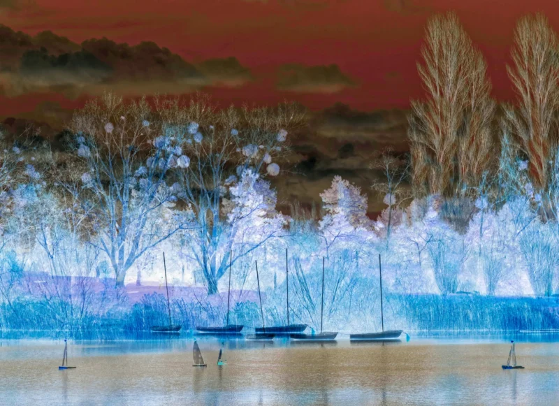 Monique · 20/07/2026 · 4730 x 3434 Cette photographie met en scène un paysage aquatique avec des bateaux amarrés en premier plan. La composition est équilibrée, avec une ligne d’horizon centrale et des arbres dénudés en arrière-plan. Le cadrage est large, capturant une étendue d’eau calme.La lumière semble artificiellement modifiée, créant un effet irisé et surréaliste. Les couleurs dominantes sont des teintes bleutées et violettées, avec des touches de rouge et de rose dans le ciel. Le style est onirique et stylisé, évoquant une ambiance mystérieuse et presque fantastique.