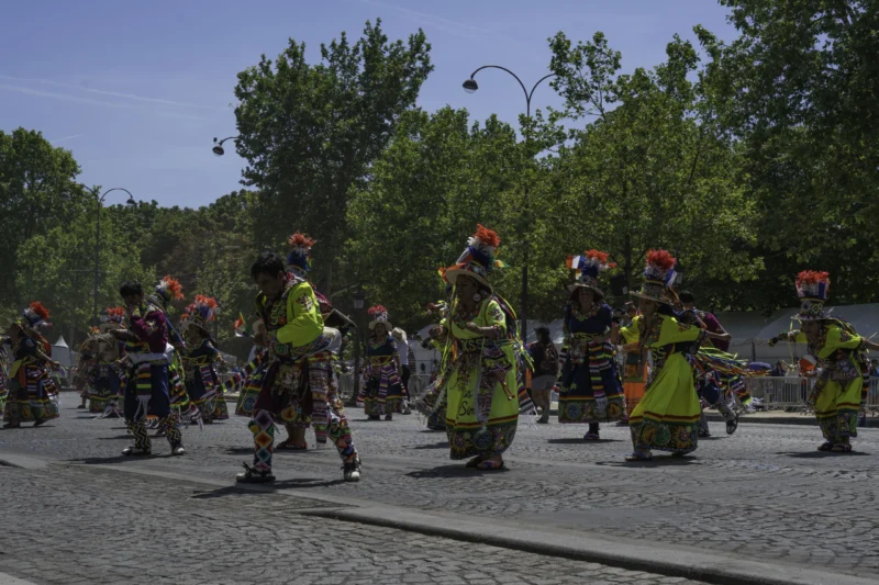 Alice · 05/07/2026 · 6072 x 4048 Cette photographie capture une parade folklorique. Le **sujet principal** est un groupe de danseurs vêtus d’uniformes colorés (vert, rouge) et coiffés de chapeaux rouges, jouant des instruments.La **composition** montre une scène dynamique en mouvement, avec un **cadrage serré** sur les danseurs en première ligne. L’**ambiance** est festive et énergique.La **lumière** est naturelle, en plein jour, avec des ombres douces. Les **couleurs dominantes** sont les verts, rouges et jaunes vifs, contrastant avec le fond plus neutre (arbres, route). Le **style** est documentaire et vibrant, reflétant une tradition culturelle.