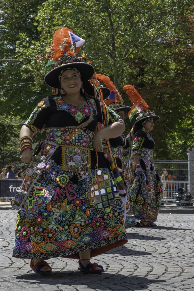 Alice · 05/07/2026 · 2468 x 3702 Cette photographie capture un groupe de personnes vêtues de costumes traditionnels colorés et ornés. Le sujet principal semble être une femme au premier plan, portant une robe multicolore et un chapeau décoré. La composition montre un cadrage serré sur les participants, avec une légère profondeur de champ.La lumière est naturelle et semble provenir d’une source externe, éclairant les détails des costumes. Les couleurs dominantes sont vives et contrastées, avec des motifs géométriques et des teintes chaudes. L’ambiance est festive et culturelle, évoquant une célébration ou une parade traditionnelle.