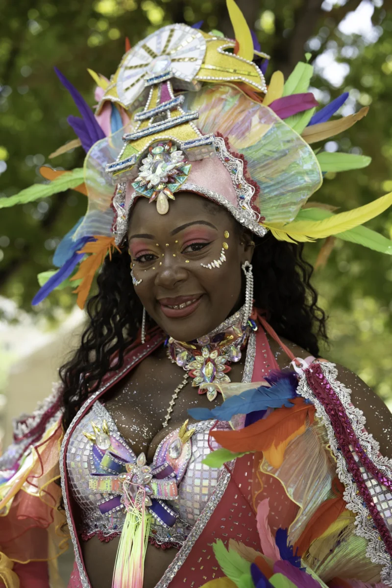 Alice · 05/07/2026 · 4672 x 7008 Cette photographie met en scène une personne vêtue d’un costume extravagant et coloré, sujet principal. La composition est centrée sur elle, avec un cadrage serré qui isole son visage et ses accessoires.La lumière semble naturelle, mettant en valeur les détails du costume. Les couleurs dominantes sont vives : roses, bleus, verts et ors, créant un contraste éclatant. Le style est festif et artistique, évoquant une célébration ou un carnaval.L’ambiance est joyeuse et exubérante, renforcée par les éléments décoratifs et la présence de la nature en arrière-plan.