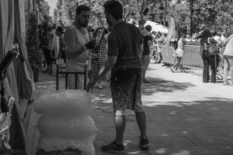 Alice · 05/07/2026 · 6506 x 4337 Cette photographie en noir et blanc capture deux hommes en interaction dans un cadre urbain. Le sujet principal est le dialogue entre eux, l’un ajustant quelque chose sur le bras de l’autre. La composition montre un espace public animé en arrière-plan, avec des passants et des éléments architecturaux.Le cadrage est serré sur les deux hommes, avec une profondeur de champ qui floute légèrement l’arrière-plan. La lumière est naturelle, contrastée, mettant en valeur les textures et les ombres. Le style est documentaire et spontané, évoquant une scène de vie quotidienne. L’ambiance est authentique et un peu mélancolique, renforcée par le monochrome.