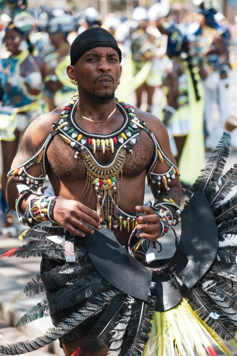 Daniel · 05/07/2026 · 3389 x 5084 Cette photographie met en scène un danseur traditionnel richement paré, sujet principal. Il porte des bijoux et des plumes, vêtu de manière traditionnelle, évoquant une culture africaine.Le cadrage est serré, centré sur lui, avec un arrière-plan flou montrant une foule en mouvement. La lumière est naturelle et met en valeur ses détails. Les couleurs dominantes sont les tons chauds (or, noir, brun) et les contrastes vifs.Le style est dynamique, presque théâtral, créant une ambiance festive et culturelle, pleine d’énergie et de tradition.