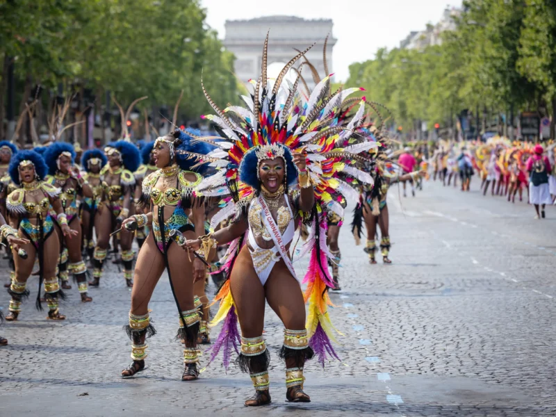 Daniel · 05/07/2026 · 4564 x 3423 Cette photographie capture une parade festive avec un danseur amérindien comme sujet principal, vêtu d’un costume richement décoré de plumes colorées et de perles. La composition montre une procession en mouvement sur une avenue pavée, avec d’autres participants en arrière-plan.Le cadrage est serré sur le danseur, mettant en valeur ses détails. La lumière naturelle, douce, éclaire la scène sans ombres marquées. Les couleurs dominantes sont vives : rouges, jaunes, bleus et verts, créant une ambiance joyeuse et dynamique. Le style évoque une célébration culturelle, tandis que l’ambiance est festive et énergique.