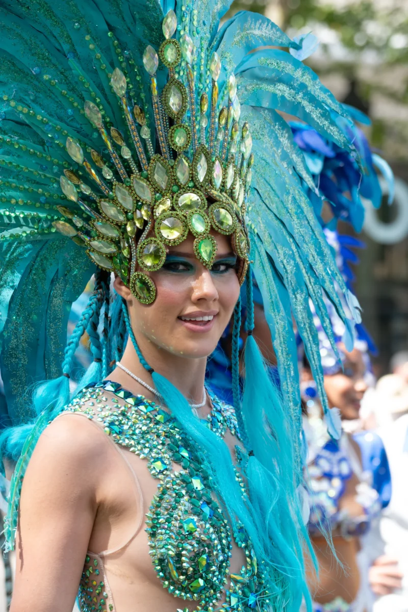 Daniel · 05/07/2026 · 3641 x 5462 Cette photographie met en scène une femme vêtue d’un costume festif et coloré. Le sujet principal est une danseuse ou une participante de défilé, portant un costume à dominantes **bleues et vertes**, avec des plumes et des paillettes.La **composition** est centrée sur elle, avec un **cadrage serré** qui isole son visage et sa tenue. La lumière est naturelle et semble douce, mettant en valeur les détails du costume. L’**ambiance** est joyeuse et festive, évoquant une célébration ou un carnaval. Les couleurs vives et le style exubérant renforcent cette impression de fête et d’énergie.