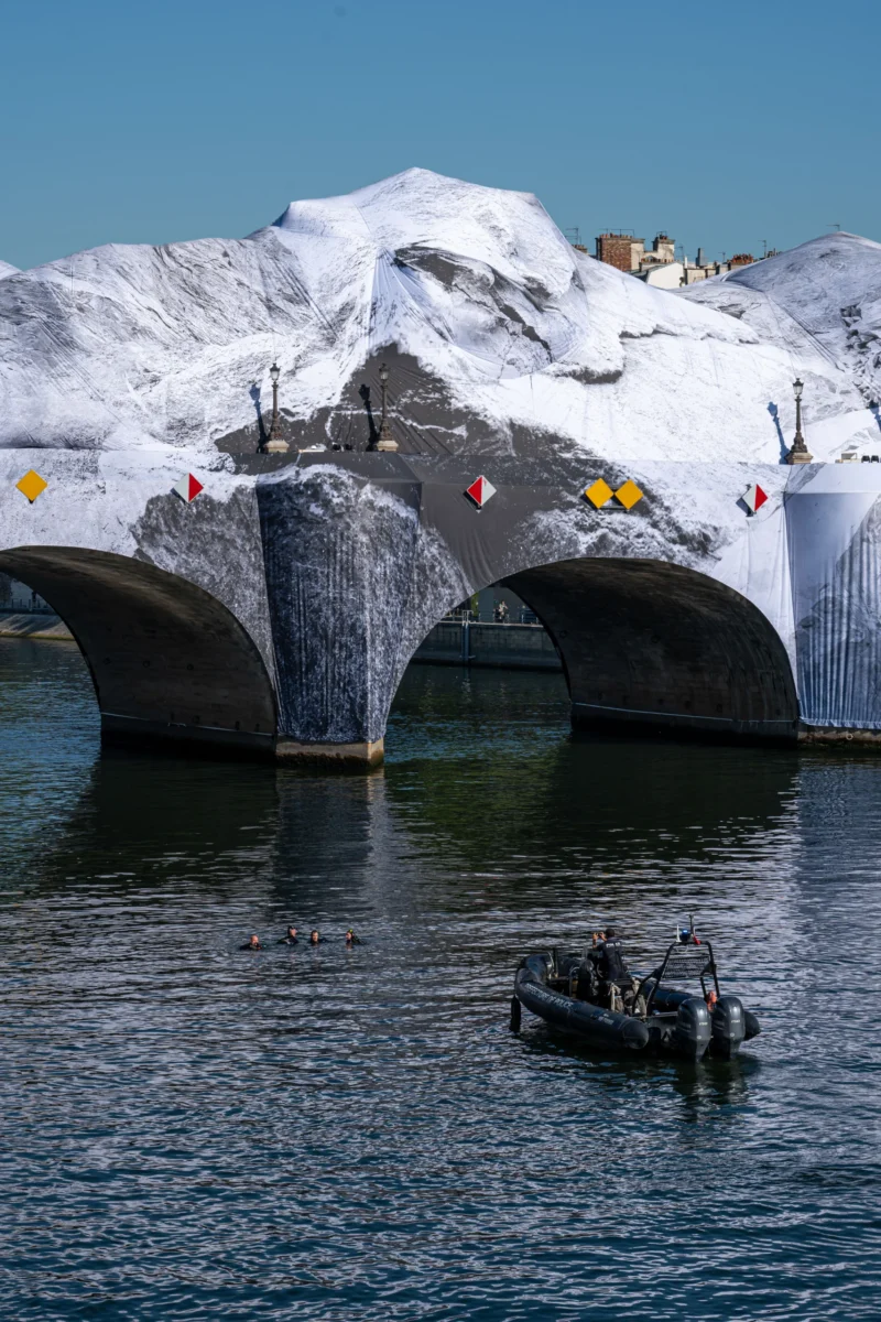Jean-Marc · 27/05/2026 · 5163 x 3442 Cette photographie met en scène un pont architectural aux arches imposantes, partiellement recouvert de neige. Le sujet principal est ce pont, avec des éléments décoratifs colorés (boules et drapeaux).La composition joue sur les contrastes entre la structure moderne et la neige, tandis que le cadrage inclut une embarcation sur l’eau en premier plan, ajoutant de la profondeur. La lumière semble naturelle, mettant en valeur les textures et les couleurs.Les couleurs dominantes sont les gris (pont), les blancs (neige) et les teintes bleutées de l’eau. Le style est photographique réaliste, avec une ambiance à la fois froide et esthétique, évoquant un lieu touristique ou artistique.