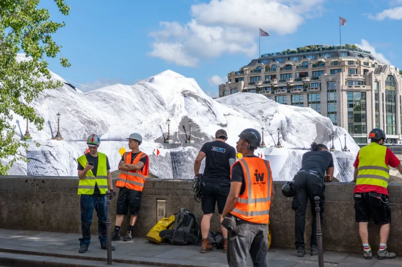 Jean-Marc · 01/07/2026 · 4206 x 2804 Cette photographie montre un groupe de personnes en tenue de travail (vêtus de gilets jaunes et orange) près d’une structure recouverte de bâches blanches. Le sujet principal semble être une intervention ou une installation urbaine.La composition est dynamique, avec un cadrage serré sur les individus et leur environnement immédiat. La lumière est naturelle, légèrement contrastée, suggérant un jour ensoleillé. Les couleurs dominantes sont les tons blancs des bâches, les jaunes et oranges des vêtements, et les gris des bâtiments en arrière-plan.Le style est documentaire, capturant une scène réaliste. L’ambiance est professionnelle et industrielle, évoquant un chantier ou une opération urbaine en cours.