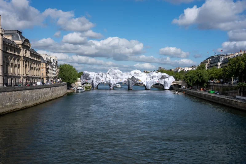 Jean-Marc · 01/07/2026 · 6048 x 4032 Cette photographie montre un pont parisien enjambant une rivière, probablement la Seine. Le sujet principal est une installation artistique éphémère composée de structures blanches en forme de coques ou de bulles, reflétées dans l’eau.La composition est symétrique, avec un cadrage large capturant les bâtiments typiques de Paris sur les deux rives. La lumière est naturelle, éclatante mais douce, avec des nuages dispersés. Les couleurs dominantes sont les tons gris-beige des bâtiments, le bleu du ciel et le blanc des installations. Le style est réaliste et photographique, créant une ambiance à la fois moderne et poétique, mêlant urbanité et art contemporain.