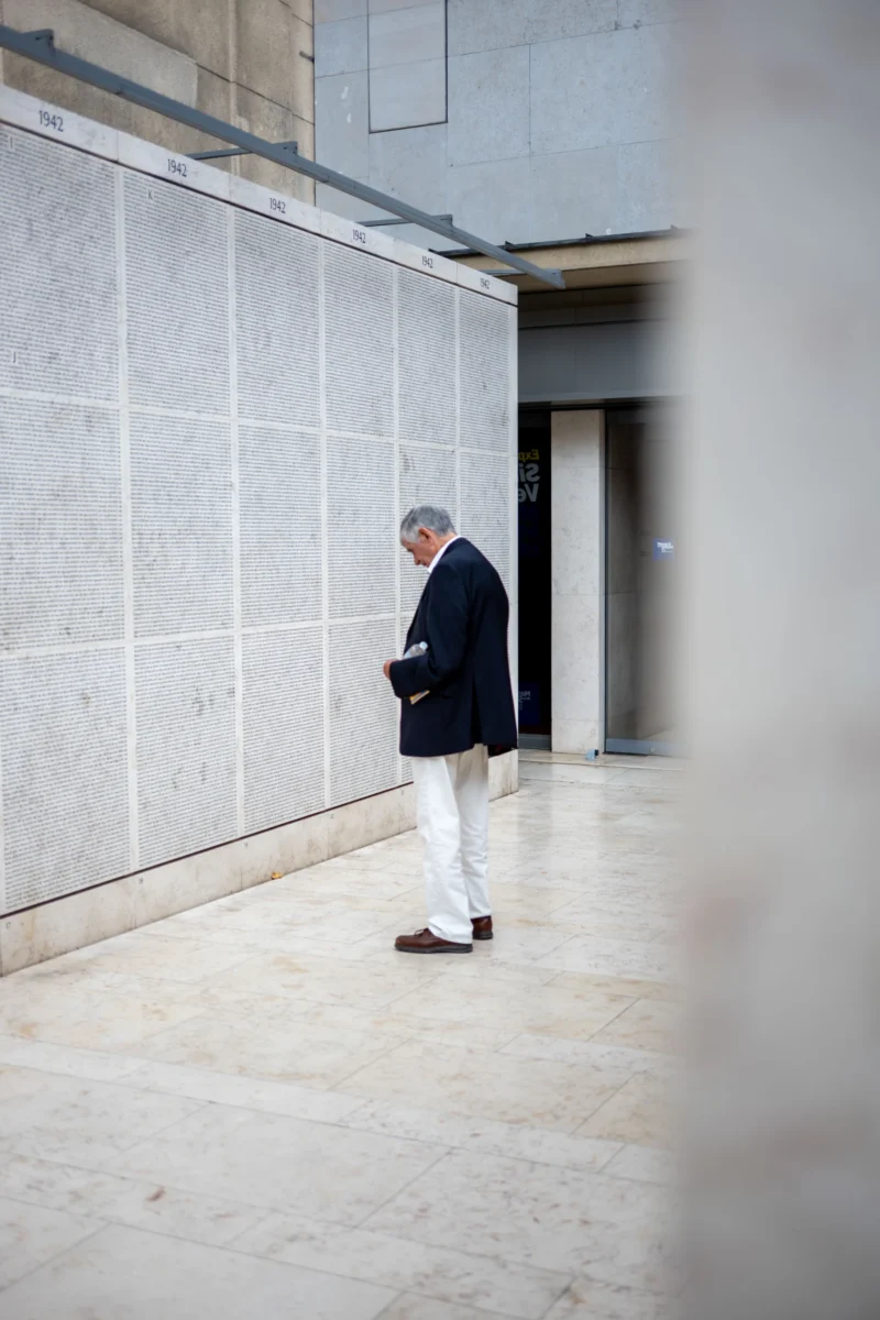 Jean-Marc · 01/07/2026 · 6048 x 4032 Cette photographie met en scène un homme âgé comme sujet principal, vêtu d’un costume sombre et d’un chapeau, marchant dans un couloir étroit. La composition est simple, axée sur une perspective linéaire avec des murs gris clairs et un sol nu.Le cadrage est serré, légèrement flou, ce qui accentue l’isolement du personnage. La lumière est naturelle, douce et tamisée, créant une ambiance sobre et mélancolique. Les couleurs dominantes sont les tons gris et beiges, renforçant une atmosphère sobre et minimaliste. Le style est épuré et réaliste, évoquant une sensation de calme et de solitude.