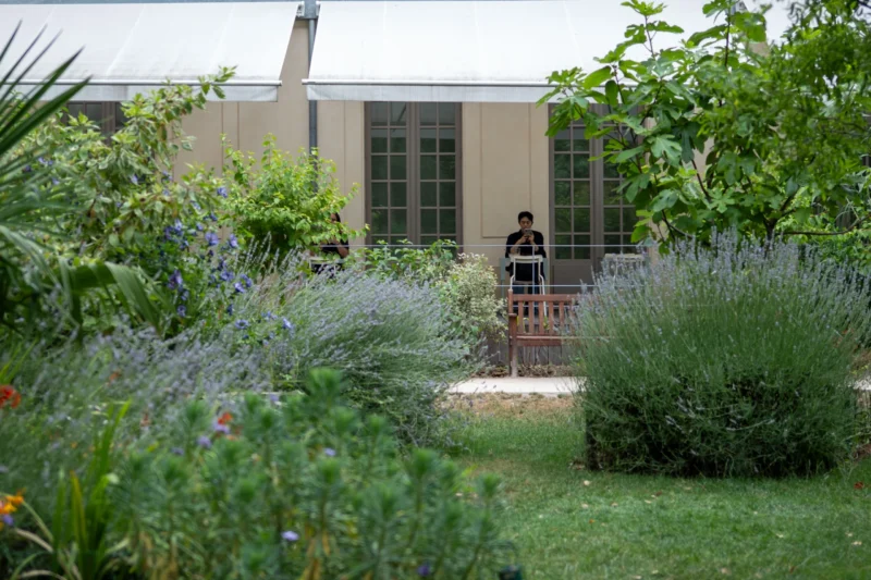 Jean-Marc · 01/07/2026 · 2328 x 3492 Cette photographie met en scène une personne assise sur un banc dans un jardin. Le sujet principal est cette silhouette, entourée d’une végétation luxuriante.La composition est équilibrée, avec un cadrage en plan moyen, mettant en valeur les plantes et le bâtiment en arrière-plan. La lumière semble naturelle, douce et légèrement tamisée, créant une ambiance paisible.Les couleurs dominantes sont les verts variés des plantes, avec des touches de gris pour le bâtiment et des nuances bleutées. Le style est réaliste et apaisant, évoquant une atmosphère calme et contemplative.