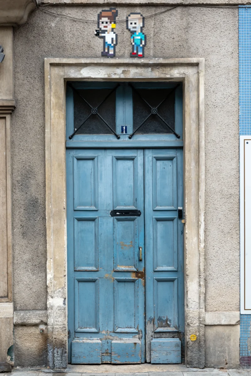 Jean-Marc · 01/07/2026 · 3945 x 2630 Cette photographie met en scène une porte bleue délavée, légèrement abîmée, encadrée par un bâtiment en pierre grise. Le sujet principal est complété par deux stickers représentant des personnages stylisés, placés au-dessus de la porte.La composition est symétrique, centrée sur la porte. Le cadrage est serré, mettant en valeur les détails architecturaux et les éléments décoratifs. La lumière semble naturelle, douce et légèrement tamisée, créant des ombres discrètes.Les couleurs dominantes sont le bleu délavé de la porte et le gris de la pierre. Le style est urbain et artistique, évoquant une ambiance à la fois vintage et moderne, avec une touche de créativité populaire.