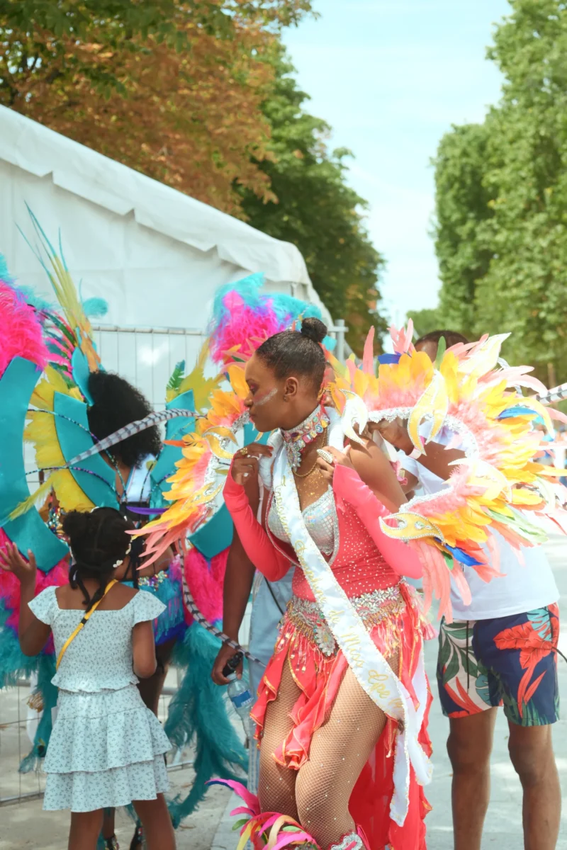 Jojo · 05/07/2026 · 3984 x 2656 Cette photographie capture une scène festive de carnaval. Le sujet principal est un danseur vêtu de plumes colorées et d’un costume rose, entouré d’enfants. La composition montre un mouvement dynamique, avec des participants en premier plan et un fond flou.Le cadrage est serré, mettant en valeur les détails des costumes et des expressions. La lumière est naturelle, légèrement tamisée, créant une ambiance joyeuse et vibrante. Les couleurs dominantes sont vives : rose, bleu, vert et jaune, typiques des célébrations carnavalesques. Le style est spontané et festif, évoquant une ambiance chaleureuse et énergique.