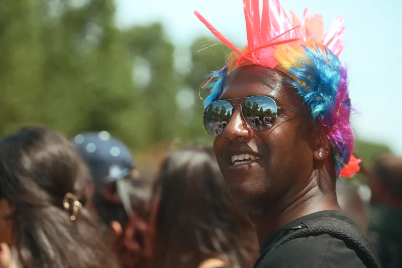 Jojo · 05/07/2026 · 3984 x 2656 Cette photographie met en scène une personne souriante, vêtue d’un haut noir, avec une coiffure colorée et extravagante en forme de crête multicolore (bleu, rose, violet). Le cadrage est serré, centré sur son visage, avec des éléments flous en arrière-plan, suggérant une foule.La lumière est naturelle et semble ensoleillée, créant des contrastes doux. Les couleurs dominantes sont vives et joyeuses, notamment les teintes pastel de la coiffure. Le style est festif et décontracté, évoquant une ambiance de célébration ou de fête en extérieur, comme un festival ou une manifestation colorée.