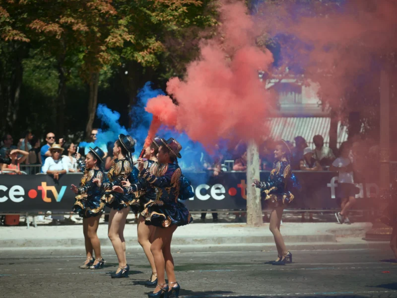 Jojo · 05/07/2026 · 5120 x 3840 Cette photographie capture une équipe de danseurs ou pompiers en tenue noire et jaune, participant à une parade. Le sujet principal est leur performance synchronisée, entourée de fumée colorée (bleue et rouge).La composition montre un cadrage dynamique, avec un premier plan mettant en valeur les danseurs. L’arrière-plan inclut des spectateurs et des véhicules décorés. La lumière est naturelle et vive, typique d’un jour ensoleillé. Les couleurs dominantes sont les teintes vives des fumigènes, contrastant avec les tenues sombres des participants.L’ambiance est festive et énergique, reflétant une célébration ou un événement public.
