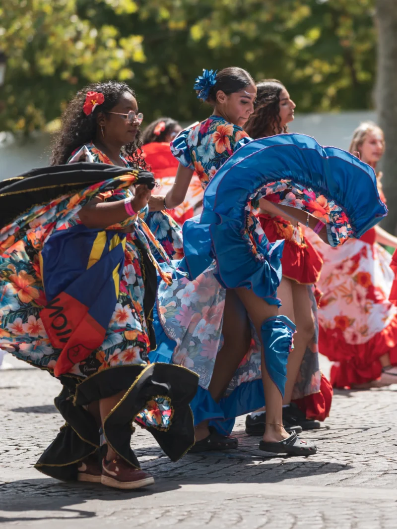 Daniel · 05/07/2026 · 4695 x 6260 Cette photographie capture un groupe de danseuses en mouvement, vêtues de robes colorées et de foulards dans les tons bleus, rouges et blancs. Le sujet principal est leur performance dynamique, suggérant une danse traditionnelle ou folklorique.La composition est serrée, centrée sur les danseuses, avec un cadrage en plan moyen. La lumière naturelle, vive et contrastée, met en valeur les couleurs vives et les détails des tenues. L’ambiance est joyeuse et énergique, évoquant une célébration ou une fête en extérieur.
