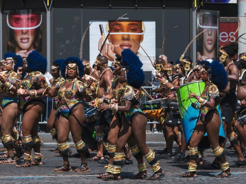 Daniel · 05/07/2026 · 3030 x 2020 Cette photographie capture une troupe de danseurs en costume traditionnel, probablement lors d’un défilé ou d’une fête. Le sujet principal est le groupe en mouvement synchronisé, vêtu de tenues colorées et ornées de plumes et d’accessoires dorés.La composition est dynamique, avec un cadrage serré sur les danseurs en premier plan. L’arrière-plan montre des spectateurs et des écrans publicitaires, créant un contraste entre tradition et modernité. La lumière est naturelle et vive, mettant en valeur les couleurs vives et chaudes des costumes. L’ambiance est festive et énergique, reflétant la vitalité et la célébration.
