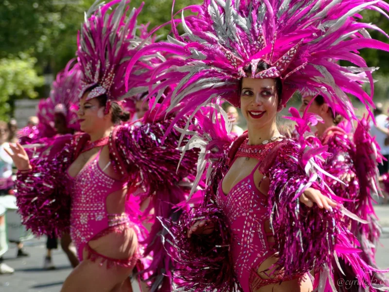 Cyrille · 05/07/2026 · 1200 x 900 Cette photographie met en scène un groupe de danseuses en costumes festifs. Le **sujet principal** est ces femmes, vêtues de tenues étincelantes à paillettes, avec des **plumes roses** imposantes.La **composition** est dynamique, avec un **cadrage serré** sur les danseuses, suggérant le mouvement. La **lumière** semble naturelle, bien que renforcée pour accentuer les reflets des paillettes. Les **couleurs dominantes** sont le **rose vif** et le **argenté**, créant une ambiance **joyeuse et festive**.Le **style** évoque un spectacle de carnaval ou une parade, tandis que l'**ambiance** est **énergique et célébratoire**.