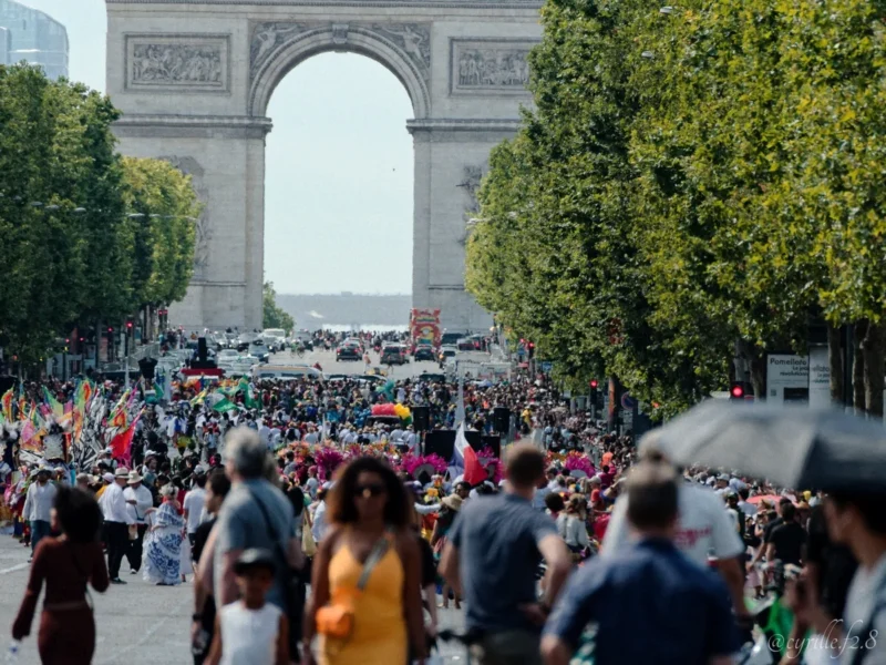 Cyrille · 05/07/2026 · 1200 x 900 Cette photographie capture l'Arc de Triomphe à Paris, sujet principal emblématique. La composition montre une foule dense en contrebas, créant un effet de profondeur vers le monument. Le cadrage est large, incluant les arbres et les véhicules en arrière-plan.La lumière est naturelle, probablement en journée, avec une ambiance lumineuse et claire. Les couleurs dominantes sont les verts des arbres, les tons neutres des bâtiments et les couleurs variées des vêtements des passants. Le style est documentaire et réaliste, reflétant une scène dynamique et animée, évoquant une ambiance urbaine vibrante et touristique.