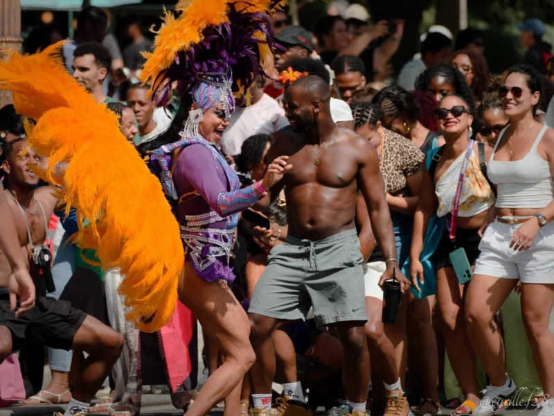 Cyrille · 05/07/2026 · 1200 x 900 Cette photographie capture une scène festive en extérieur. Le sujet principal semble être un groupe de personnes en liesse, probablement lors d’un événement carnavalesque ou une fête de rue.La composition est dynamique, avec des individus en mouvement, certains dansant ou interagissant. Le cadrage est serré, mettant en avant les visages et les expressions. La lumière est naturelle et vive, suggérant un jour ensoleillé. Les couleurs dominantes sont vives : oranges, jaunes, blancs et touches de violet, renforçant l’ambiance joyeuse et festive. Le style est spontané et documentaire, reflétant une ambiance conviviale et énergique.